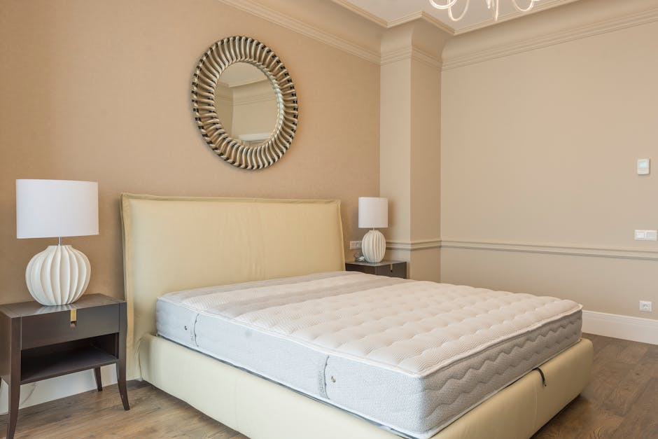 In a residential bedroom with beige walls, a large cream upholstered bed frame with a matching mattress is positioned against the wall. Two dark wooden bedside tables, each holding a white table lamp with a ribbed ceramic base, flank the bed. A round mirror with a decorative, striped frame hangs above the headboard. To the right, an open doorway leads to a hallway or another room, and on the wooden floor, part of the moving process is visible, with a partly unloaded van just outside the doorway. Furniture removal and home relocation details are implied by the setting, with a focus on the room’s interior prepared for transportation, reflecting the packing and moving services offered by Removal Companies Pimlico for house removals in Pimlico.