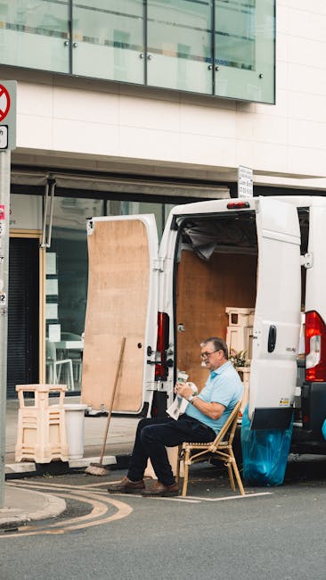 A man with glasses, wearing a light blue shirt, is seated on a wooden chair at the rear of a white delivery van parked outside a modern building with large glass windows. The van's rear doors are open, revealing bundled packing materials and cardboard boxes inside, indicating a home relocation or furniture transport process by Removal Companies Pimlico. The man appears to be taking a break during a loading or unloading activity, with a small wooden panel leaning against the van's door and additional packing structures nearby. The surrounding area features a paved street with a no-parking traffic sign visible, and the scene is lit with natural daylight, highlighting the logistics involved in house removals and packing during a moving service.