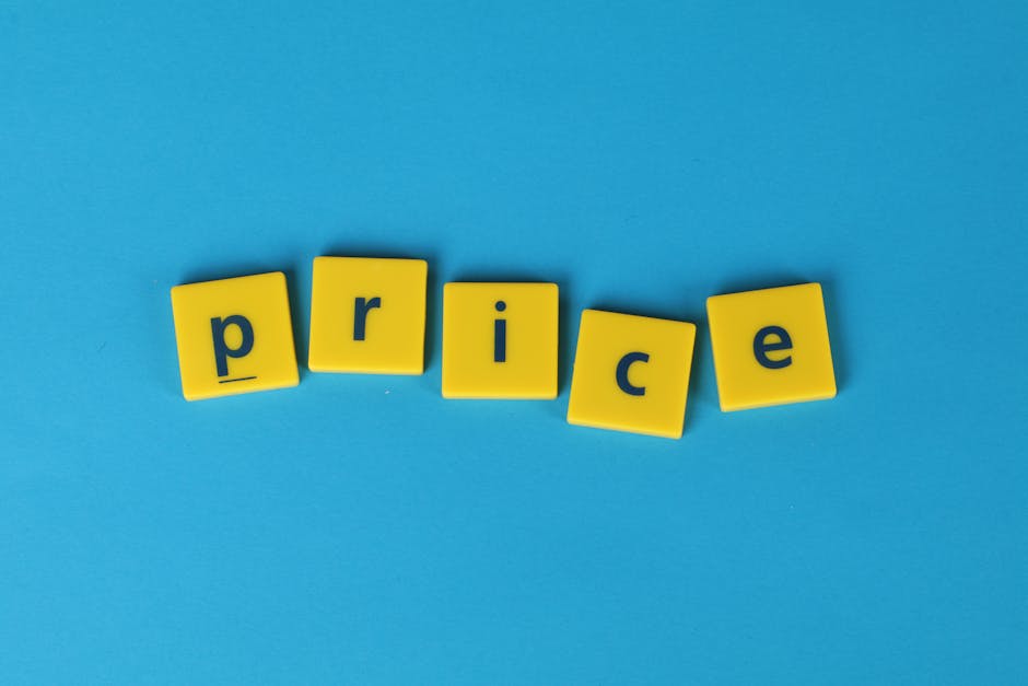 A close-up image of five yellow plastic letter blocks spelling the word 'price' laid out on a solid light blue background. The blocks are aligned in a slightly uneven row, with each block displaying a lowercase dark blue letter. In the background, there is a partially visible interior space with a wooden table or countertop and some cardboard boxes, indicating a home relocation or packing scenario associated with moving services. The lighting is natural and evenly distributed, emphasizing the vibrant colors of the blocks and background. This visual is relevant to the topic of house removals and moving logistics, as it subtly connects to the concept of pricing and service costs potentially related to removals in Pimlico, as detailed on the page about average SW1 prices, provided by Removal Companies Pimlico.