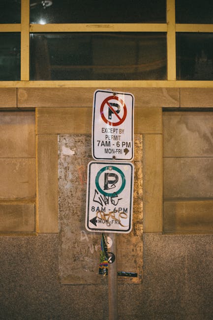 A metal parking restriction pole attached to a wall beneath a row of yellow-framed windows shows two parking restriction signs. The top sign indicates no parking except by permit from 7am to 6pm, Monday to Friday, with a red circle and slash over a letter P. The lower sign permits parking from 8am to 6pm, Monday to Friday, with a blue circle and letter P. The signs are mounted on a weathered pole that extends downward, partially obscured, and the background wall is made of stone or concrete with a textured surface, suggesting an urban environment suitable for house removals or furniture transport as facilitated by Removal Companies Pimlico.