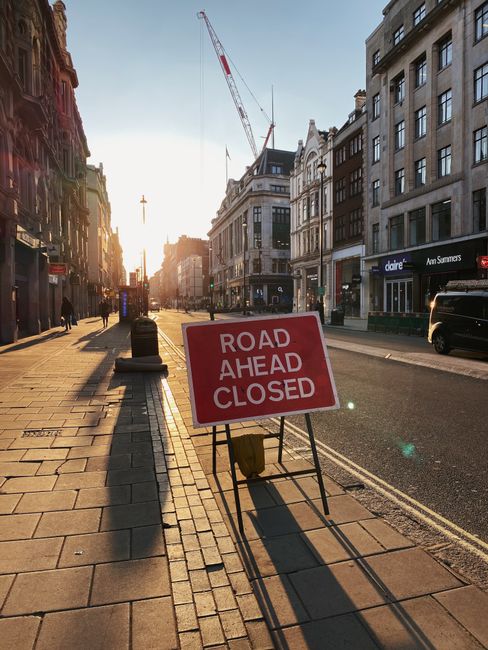 A city street during late afternoon or early evening with the sun low on the horizon, casting long shadows. The pavement is lined with commercial buildings, including a Claire store and an Ann Summers shop, both with illuminated signs. A portable metal frame holds a red and white sign reading 'ROAD AHEAD CLOSED,' placed on the sidewalk to inform pedestrians and vehicles. The sidewalk is mostly empty, with a few pedestrians walking or standing, and some outdoor seating areas visible. A construction crane extends over the rooftops, indicating nearby building work. A black van is parked along the curb, and a few bollards separate the pedestrian walkway from the road. The scene captures an active urban environment focused on building maintenance or road closure arrangements, with natural lighting highlighting details of the furniture, signage, and cityscape, reflecting typical house removals and transport logistics in Pimlico.