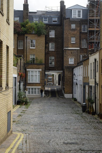 A narrow street with cobblestone pavement lined by closely spaced, multi-storey brick buildings typical of an urban residential area. The buildings feature a mix of traditional and modern architectural styles, with some windows adorned with flower boxes and others with scaffolding indicating ongoing maintenance or renovation work. At the end of the street, there is a large, older brick building with a white door and several windows, and greenery such as bushes or small trees is visible on the rooftop garden of the building. A person is standing near the entrance of the alley, and a black van is partially visible parked at the far end, suggesting the logistics involved in furniture transport and home relocation. This setting reflects an environment where house removals by companies like Removal Companies Pimlico are often required to navigate narrow streets, manage loading and unloading of packing materials such as cardboard boxes and furniture, and carefully execute loading processes in constrained spaces.
