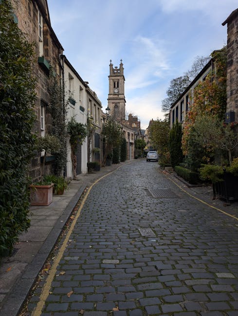 A narrow cobblestone street in Pimlico with closely spaced residential buildings on either side, featuring small front gardens and potted plants. A white van is parked further down the street, near the entrance of a property. In the background, the tall clock tower of Tate Britain is visible against a partly cloudy sky. The scene suggests a quiet urban environment suitable for home relocation or furniture transport, with the street’s limited width requiring careful planning during loading and unloading. As part of their property moving services, Removal Companies Pimlico likely coordinate the loading process using equipment such as trolleys and blankets to protect furniture and ensure smooth handling amidst the residential setting.