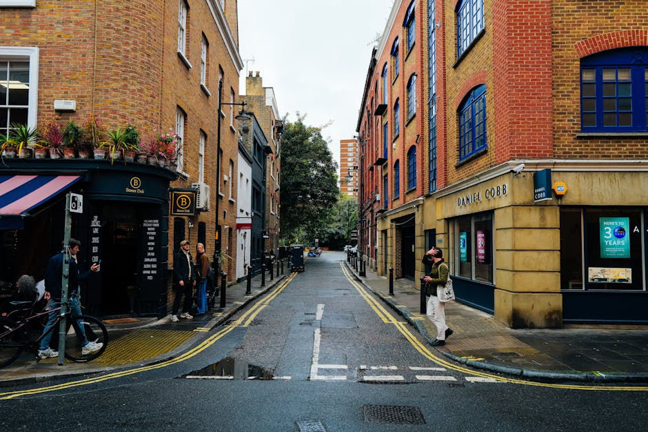 A narrow street with cobblestone pavement lined by closely spaced, multi-storey brick buildings typical of an urban residential area. The buildings feature a mix of traditional and modern architectural styles, with some windows adorned with flower boxes and others with scaffolding indicating ongoing maintenance or renovation work. At the end of the street, there is a large, older brick building with a white door and several windows, and greenery such as bushes or small trees is visible on the rooftop garden of the building. A person is standing near the entrance of the alley, and a black van is partially visible parked at the far end, suggesting the logistics involved in furniture transport and home relocation. This setting reflects an environment where house removals by companies like Removal Companies Pimlico are often required to navigate narrow streets, manage loading and unloading of packing materials such as cardboard boxes and furniture, and carefully execute loading processes in constrained spaces.