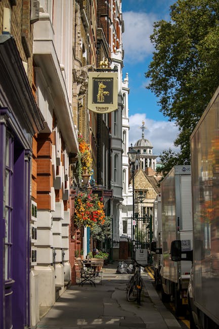 A narrow street scene in Pimlico with a row of traditional brick and stucco buildings featuring decorative facades, flower boxes, and hanging signs, including one for a restaurant named 'Young's' with a yellow lion logo, set against a blue sky with scattered clouds and a church with a dome and spire visible in the background. On the right side of the image, two large white moving vans are parked on the pavement, partially blocking the sidewalk. The vans are being used for home relocation activities, with one showing an open rear door and packing materials inside, while the other is closed, ready for transport. A small table with a chair is placed outside one of the buildings, and a bicycle is secured to a railing nearby. The scene depicts a data collection of furniture, boxes, and moving equipment as part of the loading process by Removal Companies Pimlico, illustrating the logistics involved in local house removals and furniture transport in the area.