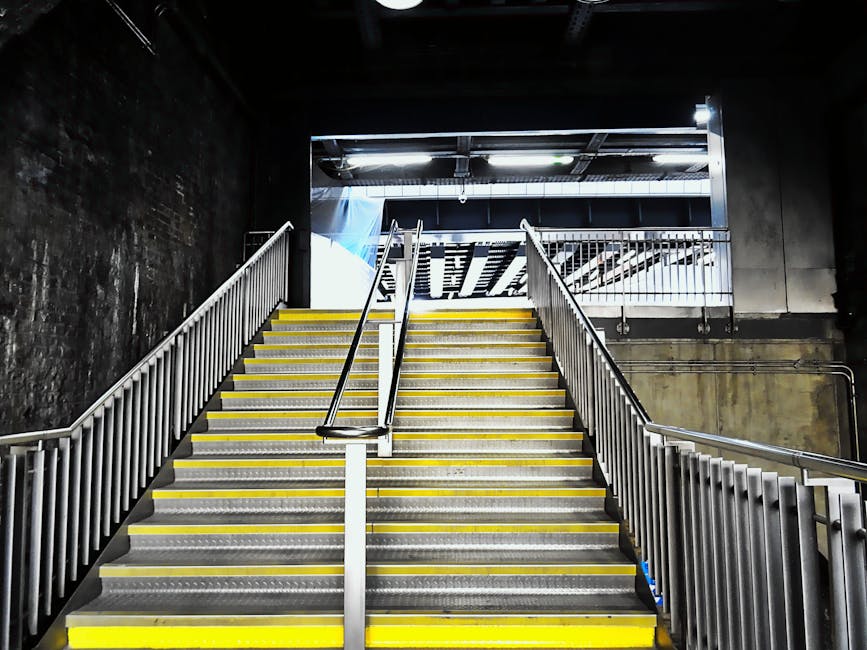 Image depicting a set of indoor stairs with metallic steps highlighted by yellow safety strips along each edge, located in a building with dark walls and overhead lighting. The staircase has a central handrail and side railings, with a metal opening at the top leading to a higher level. The surrounding environment suggests a commercial or residential property, illustrating a typical staircase that might pose access challenges during home relocation or furniture transport. This scene reflects potential staircase and lift access issues encountered during removal and moving services, where careful handling of furniture and boxes is necessary. The setting supports the logistical considerations of loading and unloading heavy or bulky items in tight or constrained spaces, as coordinated by companies like Removal Companies Pimlico.