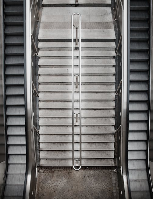 A top-down view of a concrete staircase with a central metal handrail, situated between two escalators in an indoor setting. The stairs are made of light grey concrete with visible tread lines, leading downward from a tiled floor at the top. Flanking the staircase are escalators with metal steps, each edged with black rubber or plastic, supporting a moving walkway system. The lighting is neutral, casting even illumination across the scene, which appears to be part of a commercial or public building. The environment suggests a location where efficient movement of people and, potentially, household items during a home relocation is assisted by the staircase and escalator system, noting the importance of handling furniture carefully when navigating staircase and lift access issues. As a professional removals company, Removal Companies Pimlico may assist clients in planning safe furniture transport through such areas during moving days.