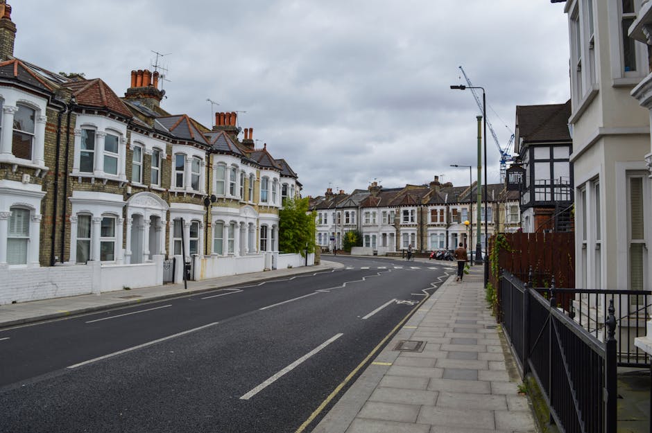 A quiet residential street in Pimlico with a row of white Victorian terraced houses featuring bay windows and brick chimneys, lined with a paved sidewalk and black metal fencing. The street is empty except for a person walking away in the distance near the curb. Overhead, a cloudy sky casts diffuse light across the scene. Visible on the right side is the edge of a modern white building with large windows. In the background, a construction crane is seen behind the homes, indicating ongoing development. The scene captures a typical day during a home relocation process, with no moving activity directly visible but implying a tranquil environment suitable for furniture transport and packing during removals, as handled by Removal Companies Pimlico.