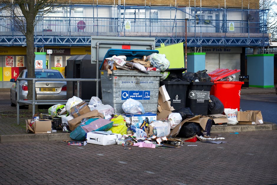 An overflowing waste disposal area in Pimlico, showing multiple rubbish bins, including a grey mixed paper and card bin, a black recycling bin, and a red general waste bin, all filled with overflowing trash such as cardboard boxes, plastic bags, paper packaging, and discarded packaging materials. The bins are situated on a paved sidewalk next to a metal railing, with some debris and paper scattered on the ground. Behind the waste area, there are commercial buildings with closed shutters and signage, and a parked car is visible to the left. The scene is outdoors, daytime, with natural light and shadows cast around the bins, illustrating the importance of proper waste disposal following local laws for house removals or relocation services, as promoted by Removal Companies Pimlico.