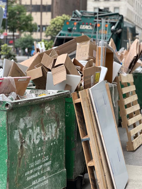 An overflowing waste disposal area in Pimlico, showing multiple rubbish bins, including a grey mixed paper and card bin, a black recycling bin, and a red general waste bin, all filled with overflowing trash such as cardboard boxes, plastic bags, paper packaging, and discarded packaging materials. The bins are situated on a paved sidewalk next to a metal railing, with some debris and paper scattered on the ground. Behind the waste area, there are commercial buildings with closed shutters and signage, and a parked car is visible to the left. The scene is outdoors, daytime, with natural light and shadows cast around the bins, illustrating the importance of proper waste disposal following local laws for house removals or relocation services, as promoted by Removal Companies Pimlico.